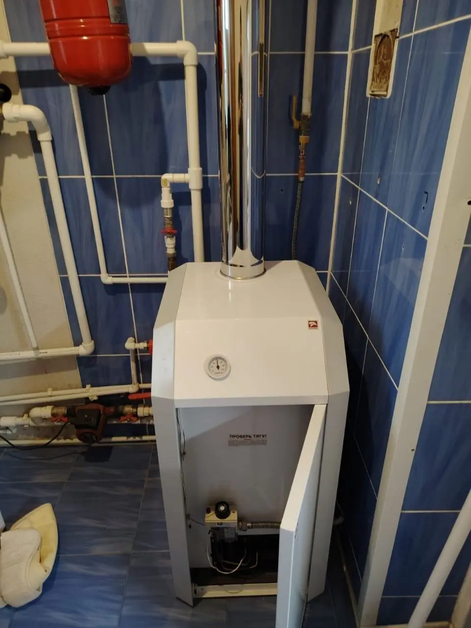 напольный газовый котёл с открытой фронтальной дверцей, видна верхняя панель с манометром, вертикальный дымоход и красный расширительный бак
